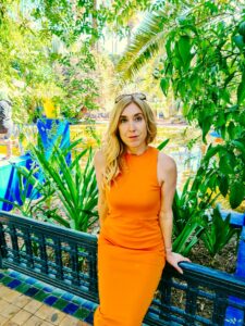 Wearing an orange dress that demonstrates style tips for women traveling to Morocco, in front of green trees in Jardin Majorelle. 